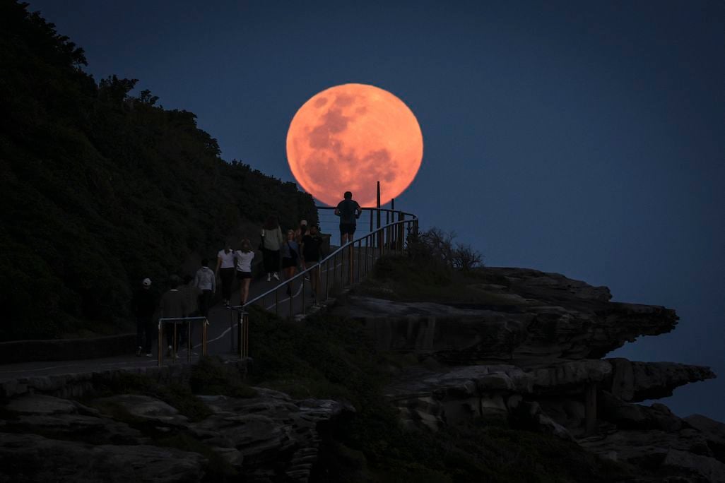 a supermoon rises and appears orange in front of a hillside near the coast, its light silhouetting a person climbing a staircase