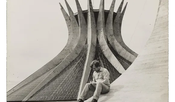Grayscale image of Robert Hughes sitting on the sloping curve of a building examining something in his hands, with a modernist church in the background.