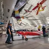 Inside a large hangar filled with natural light, three white people maneuver a small red, white, and blue airplane into its display space. At least eight aircraft are suspended from the hangar's ceiling.