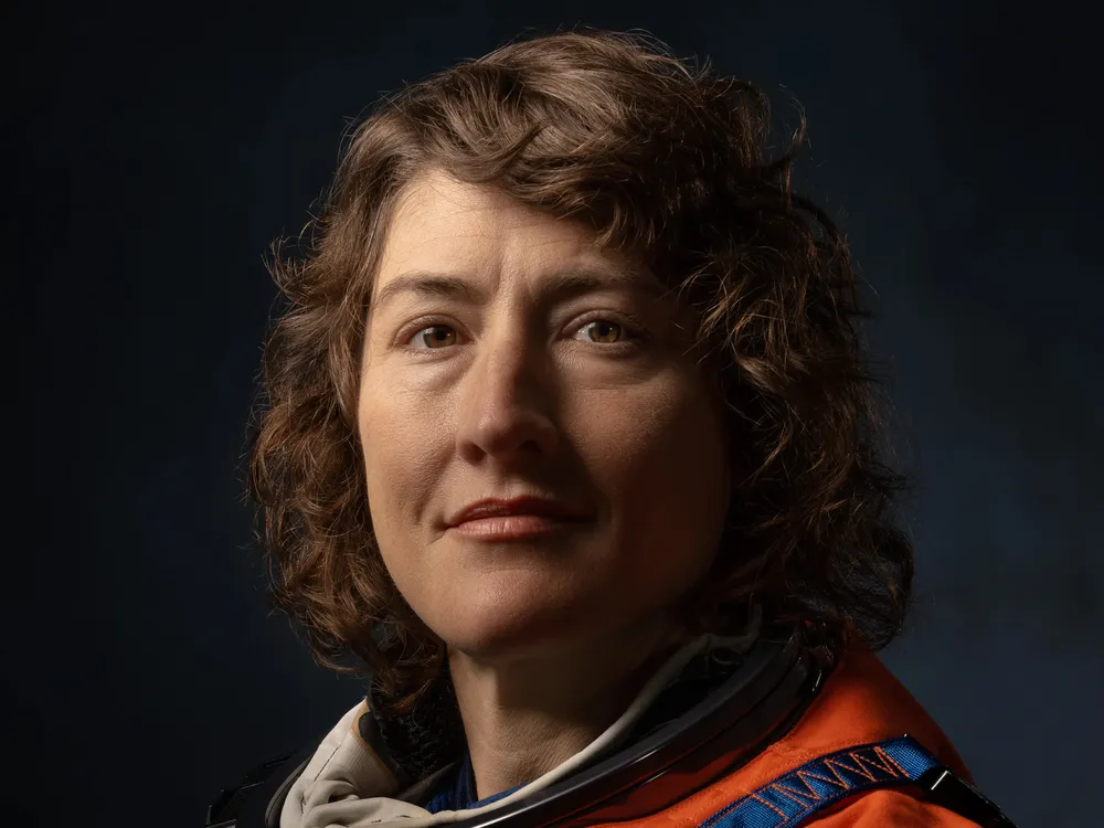 Dramatically-shot portrait of a white female astronaut in an orange spacesuit.