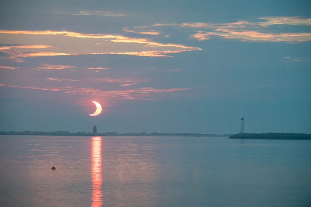 a partially eclipsed sun appears as a crescent over a lighthouse and water in early morning hours