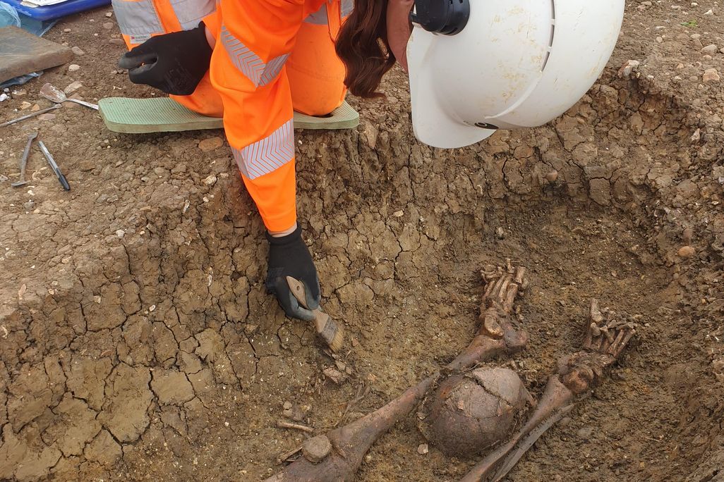 remains of skeleton partially revealed in soil, a worker in orange safety gear digging next to it
