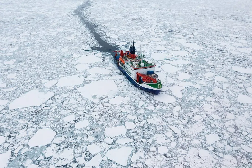 A photograph shows a white and red ship sailing through cracked sea ice 