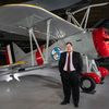 A white man in his 30s stands in front of a restored biplane displayed on a polished concrete floor inside a museum. He is wearing glasses and a dark suit with a white shirt and burgundy tie.