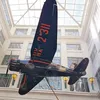 An upward-looking shot shows a dark-colored, single-engine Stinson Reliant monoplane with red markings on its wing, suspended indoors from the ceiling of a multi-story building with a glass atrium roof.