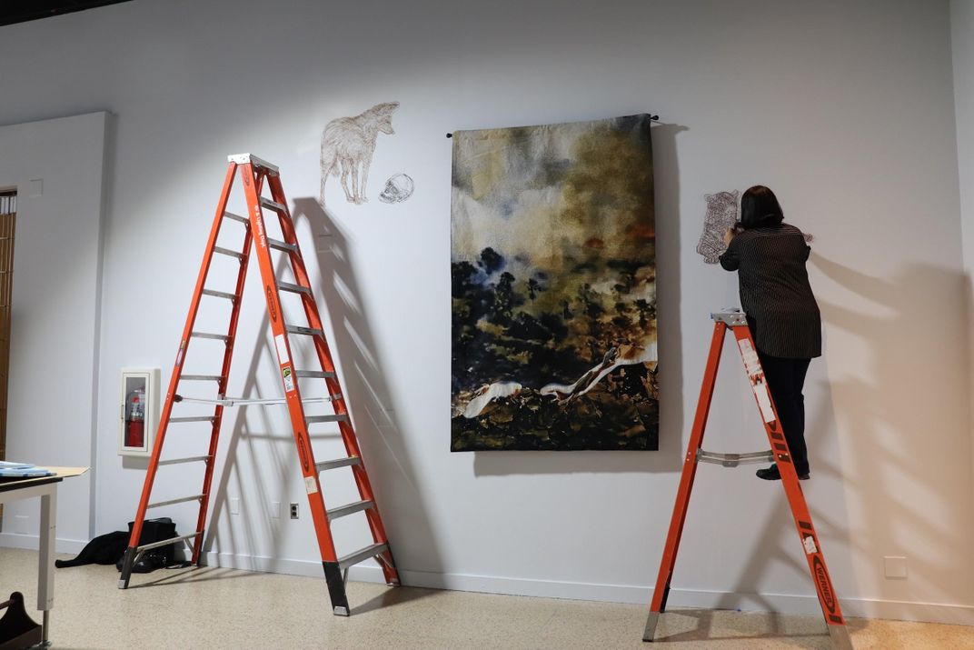 Artist on a ladder nailing string art to a white wall next to a tapestry