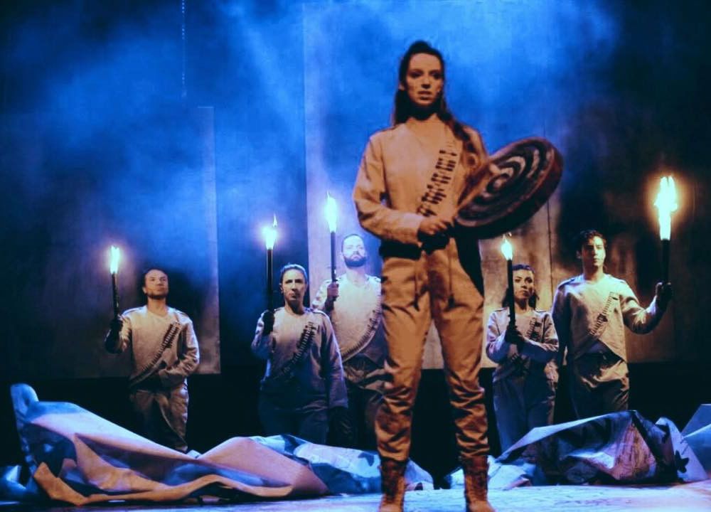 A woman stands on stage, holding a hand drum and a beater stick. Five actors behind her hold up torches. They all wearing matching white outfits with mock bandoliers. The stage is foggy and lit blue.