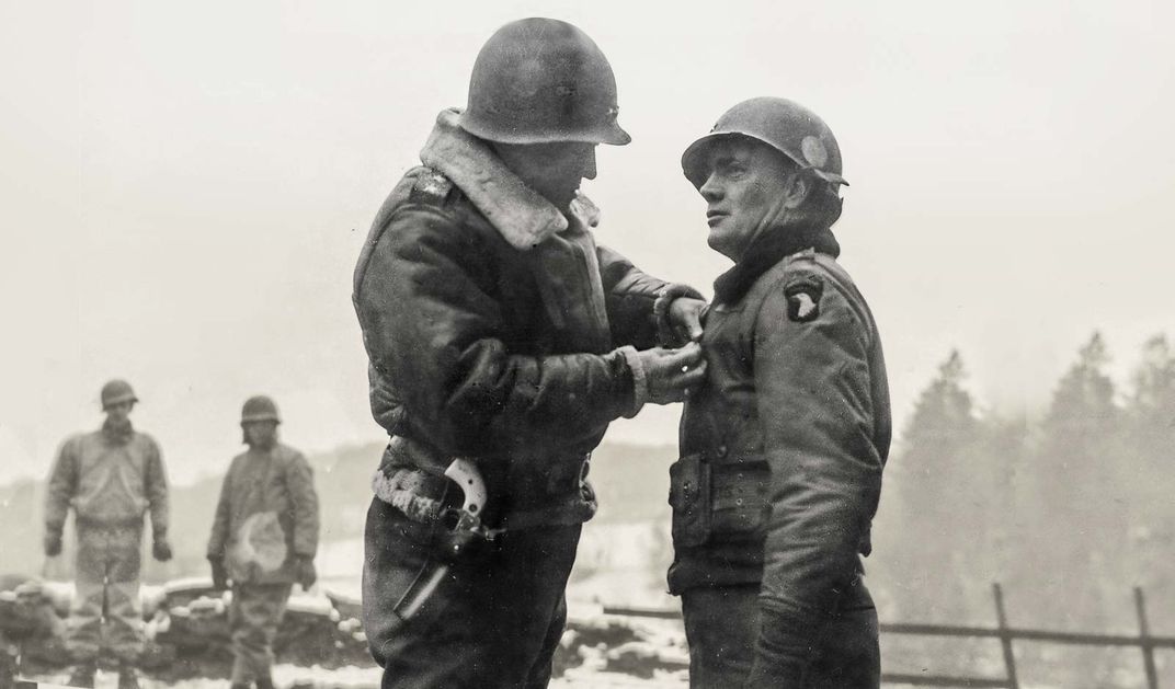 One man putting a pin on another man's jacket, both dressed in military uniforms