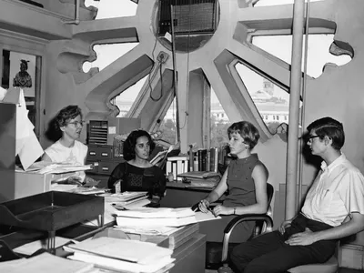 In a black and white photo, three women and one man sit together in a room with a large circular window behind them, surrounded by papers.