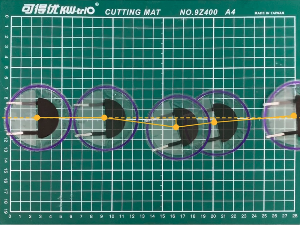 The robot jumps across a mat. It is clear with a blue ring on its rim and black and silver objects on the inside. In this time lapse, the robot jumps across the mat in a straight line.