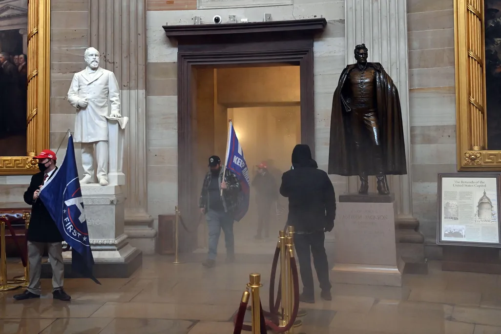 Two men carrying large blue flags - the first words of KEEP AMERICA GREAT appear in one - walk into the corridor flanked by two statues of men and huge Trumbull paintings in gilded frames, surrounded by hazy gas on the floor
