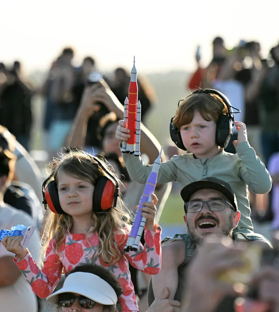 children on adults' shoulders watch the launch with small toy rockets and ear protection