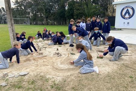 Students cluster in groups and individually around a sandy area, adding and refining the community map they are building out of sand, leaves, and sticks.