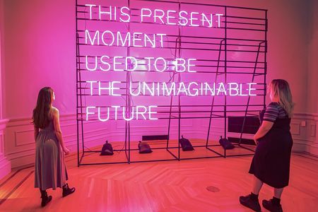 Two people stand in front of a pink neon sign that states "This Present Moment Used to Be the Unimaginable Future"