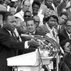 Martin Luther King, Jr. stands at a podium with several microphones. Behind him to his left is a crowd of seated and standing people, including Dorothy Height, turned towards him.