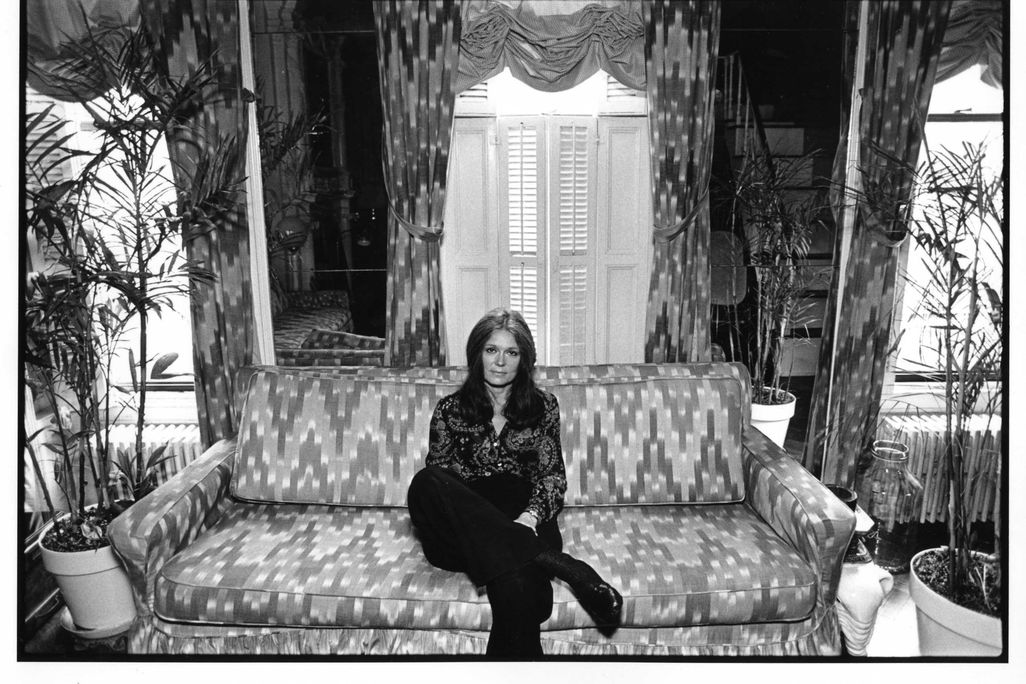 A black and white image of Steinem, a white woman in a patterned button down and black pants, sitting on a patterned couch in front of house plants and tall windows with curtains