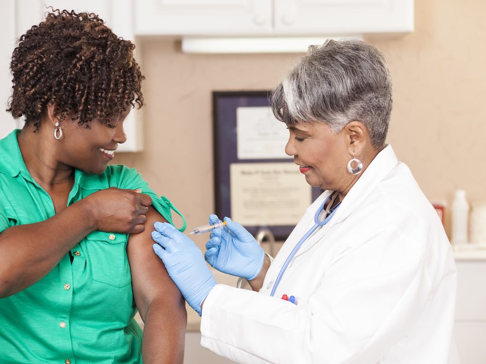 An image of a woman of color recieving a vaccine from a doctor. The doctor is a woman of color dressed in a white lab coat.