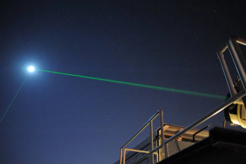 A dark night sky with some metal equipment and beams in the foreground, and then a bright green line shooting through the sky toward a bright full white moon