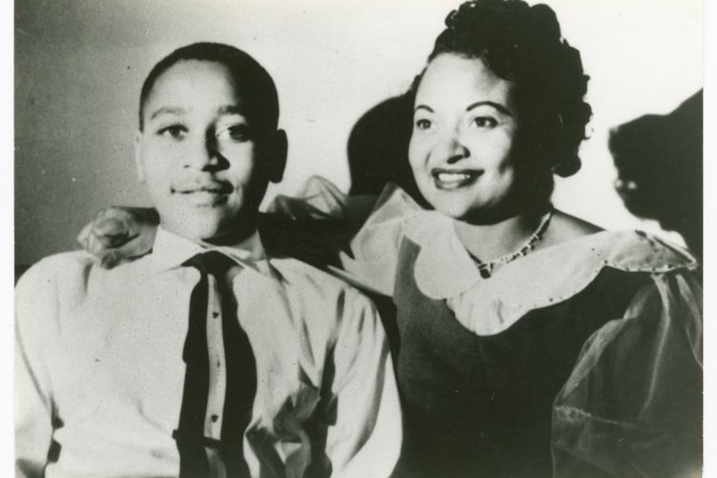 Emmett Till, a young boy in a suit and tie, smiles next to his mother, who wears a dress and has her hand on his shoulder