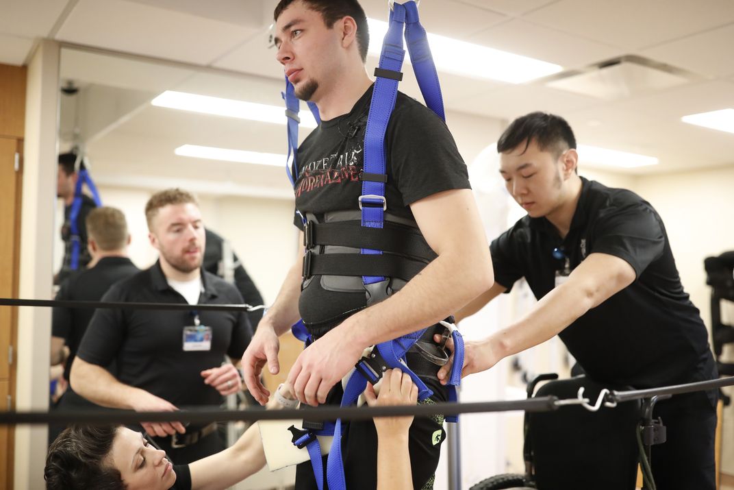 A person recovering from a spinal cord injury is strapped into a harness while standing on a treadmill.
