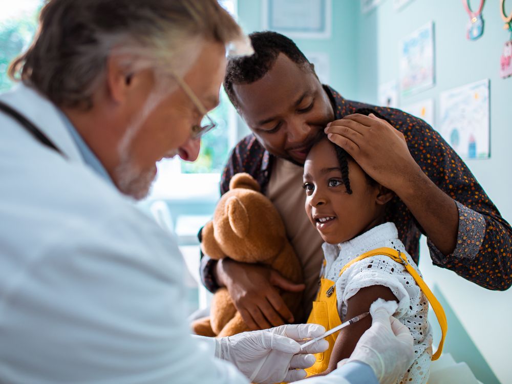 An image of a older male physican vaccinating a young child. The child is smiling and is being held by her father.