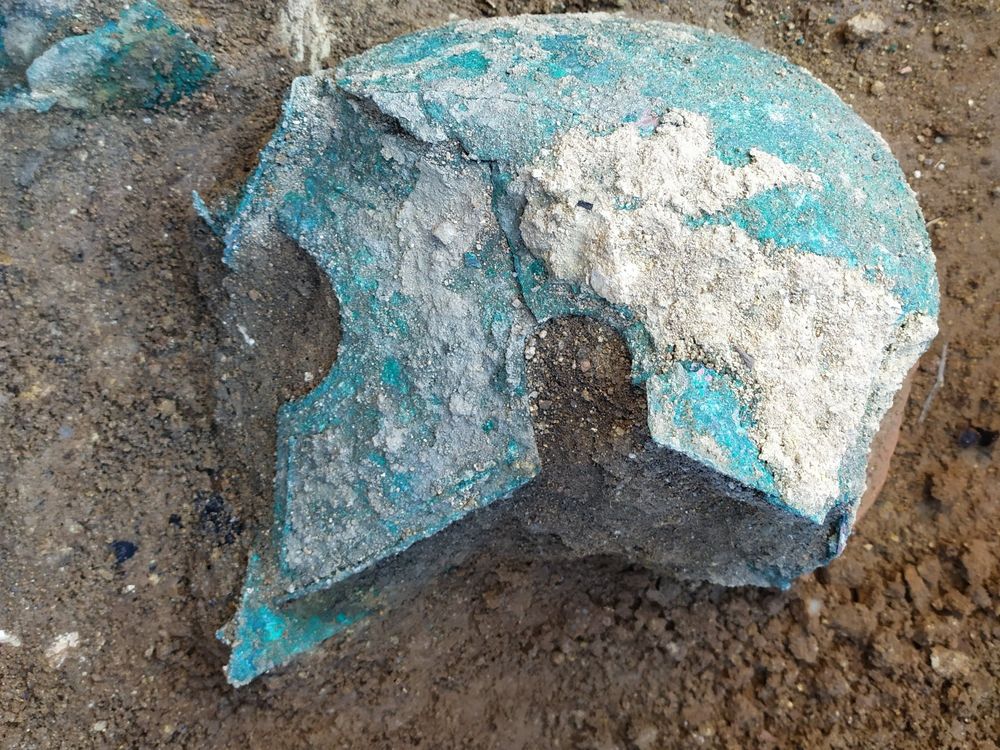 A view of a blue-green oxidized metal helmet, with sharp curved cheek plates that extend past the face, lying on the ground in the dirt