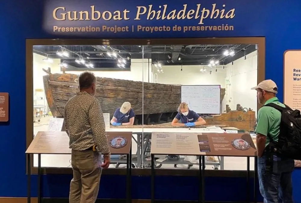 Visitors look into an exhibit window and see staff working on the wooden Gunboat