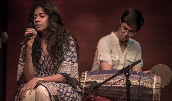 On the left, a woman with long wavy dark hair and patterned blue blouse sings into a microphone, seated, eyes closed. On the right, another person sits behind a long barrel-shaped drum, holding a hand drum, looking down.
