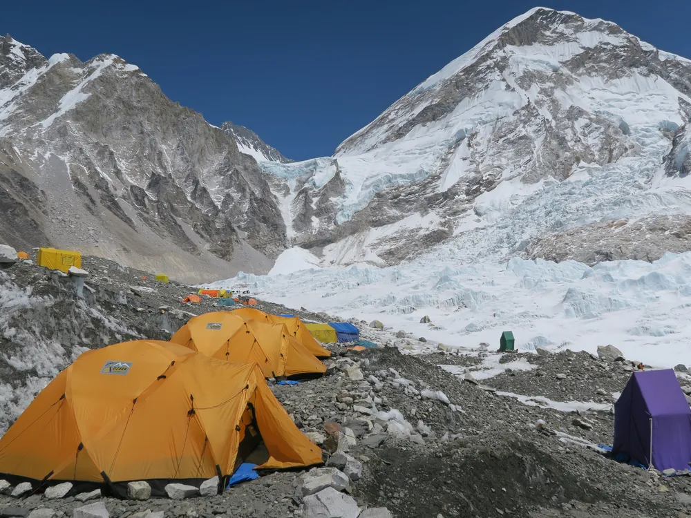 A photo of Everest Base Camp. Tents of various colors are scattered across the gray, rocky ground. Snow-covered mountains are in the background.