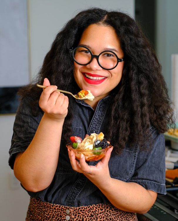 Image shows a smiling woman eating a bowl of fruit.