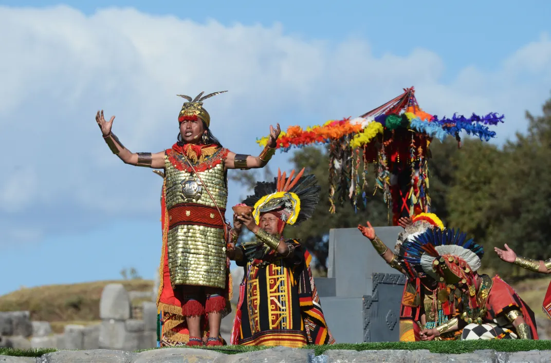 A man stands with arms raised, wearing a shiny golden outfit, a golden feathered cap, and a golden breastplate. Behind, rainbow-colored feathers and two people kneeling and raising their hands toward him