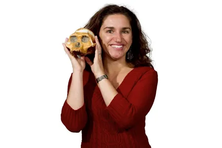 Headshot of paleoanthropologist Briana Pobiner in a red sweater holding an early human skull up text to her face on white background.
