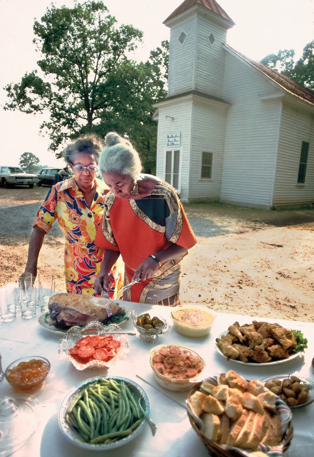 two Black women in colorful outfits plate food at an outdoor dinner with a white painted church with steeple in the background