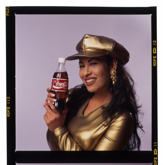 Selena in a gold cap holding a Coca-Cola bottle
