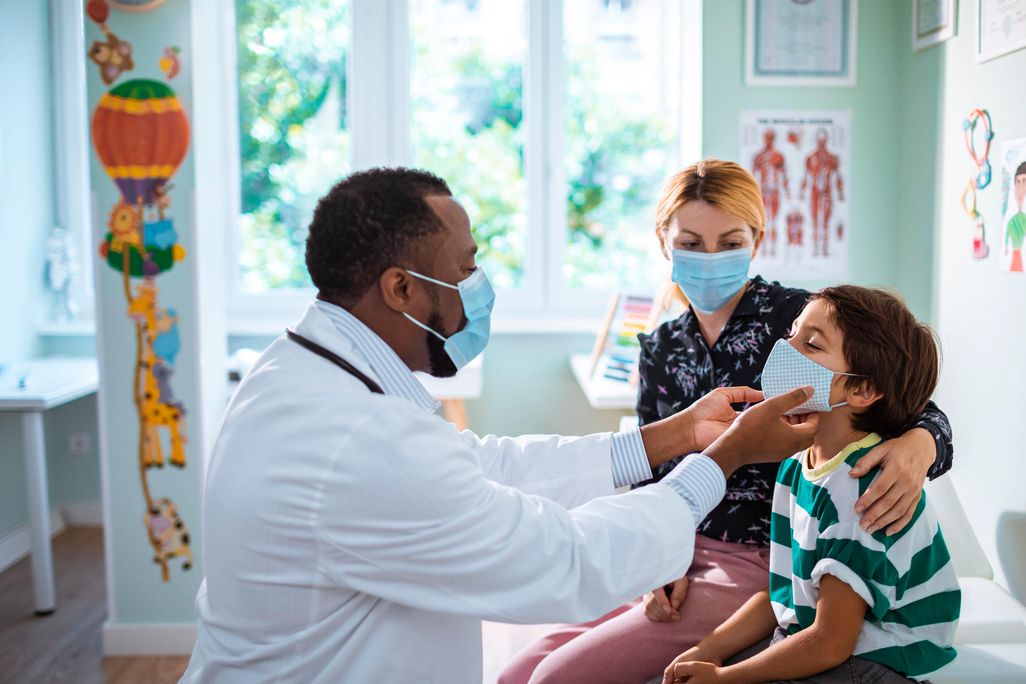 A child sits next to a woman with blond hair while a pediatrician looks at his face. All three are wearing masks.