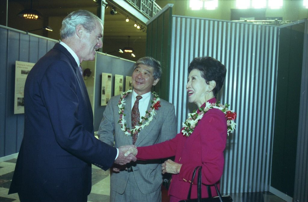 Franklin Odo stands between a man and a woman shaking hands. Odo is wearing a grey suit and flower lei.