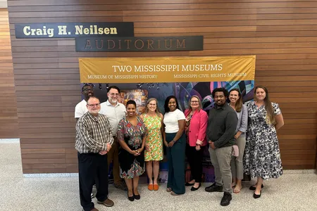 10 museum and education professionals dressed in business attire smiling and standing in front of a multicolored backdrop that reads, “Two Mississippi Museums, Museum of Mississippi History, Mississippi Civil Rights Museum”.