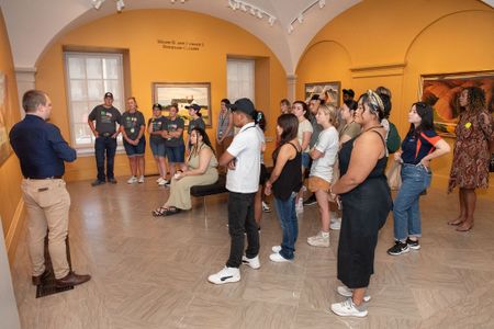 A man in a navy blue shirt and khakis leads a large group of students through an art gallery