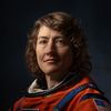 Dramatically-shot portrait of a white female astronaut in an orange spacesuit.