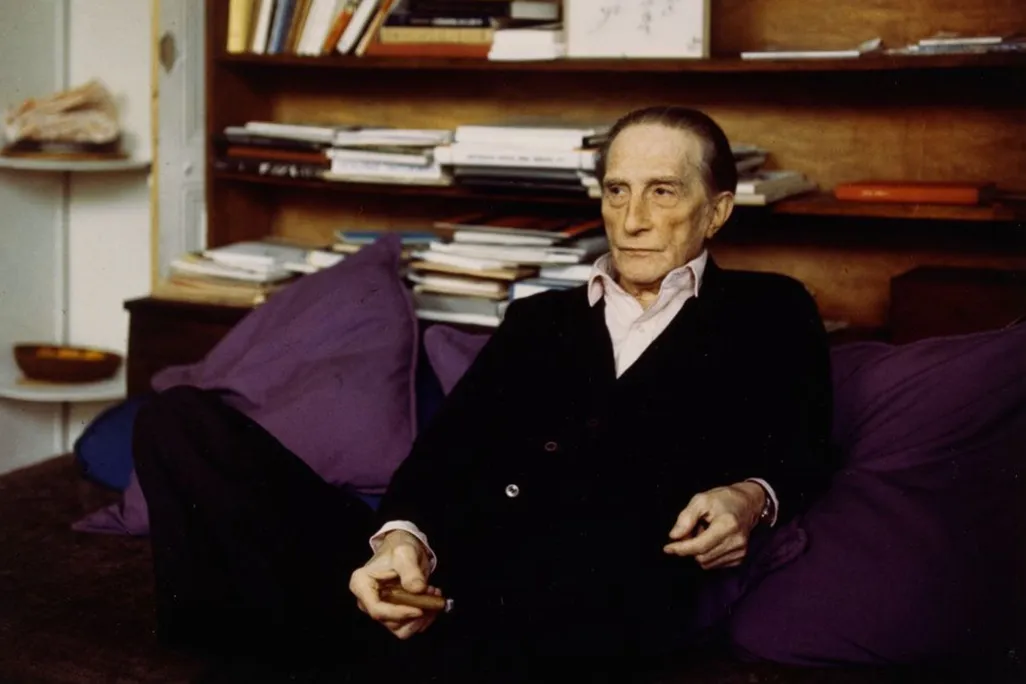 Elderly man in black sweater sitting in purple chair in front of wooden bookshelf