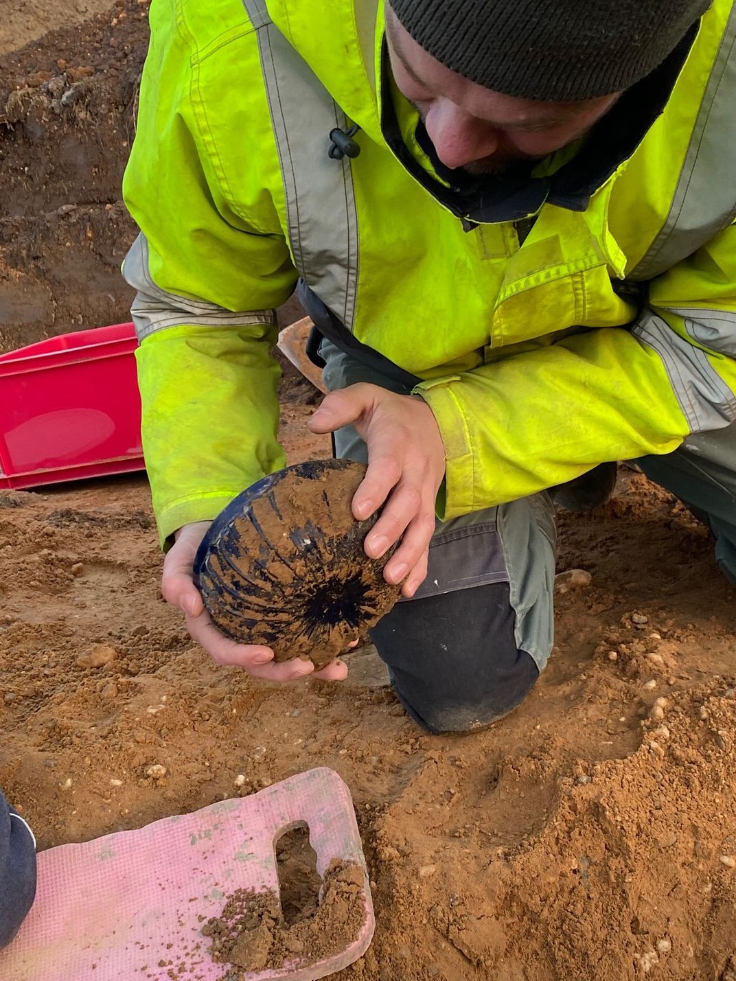 A person in a bright yellow reflective vest holds the bowl in their hands, which is covered in reddish brown dirt