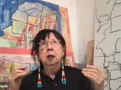 Still from a video of Jaune Quick-to-See Smith. Smith wears a black shirt with her hair pulled back, gold wire glasses, and long beaded earrings. She is sitting in front of works of art depicting maps of the United States, several in color and one in blac
