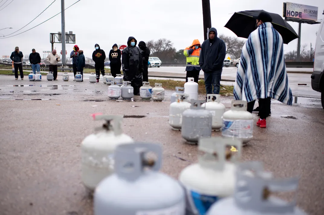 A line of people standing next to propane tanks on asphalt with puddles