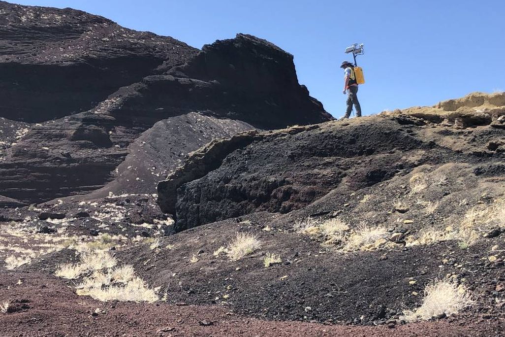 An image of a planetary scientist walking in a mountainous region with a yellow backpack on. The backpack has a camera attached to it.