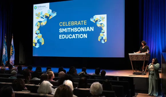 A woman stands at a podium in a theater with "Celebrate Smithsonian Education" projected on the screen