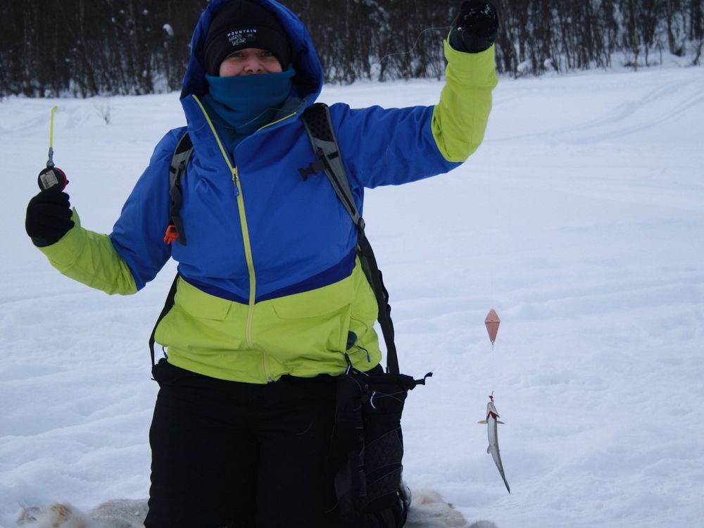 Person wearing winter gear kneeling in the snow holding up a fishing line with a small fish hooked on it