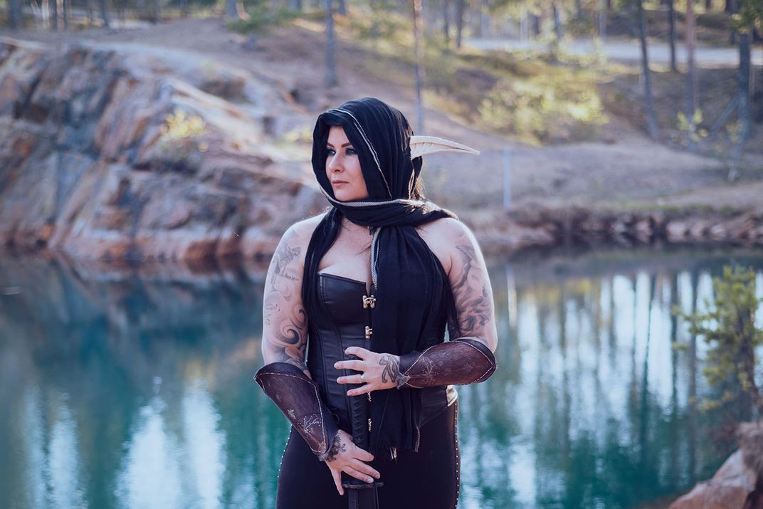 A woman poses in front of a mountain lake wearing black corset and headscarf, leather arm guards, and long, curved, pointy ears. Her bare arms show painterly tattoos in black.