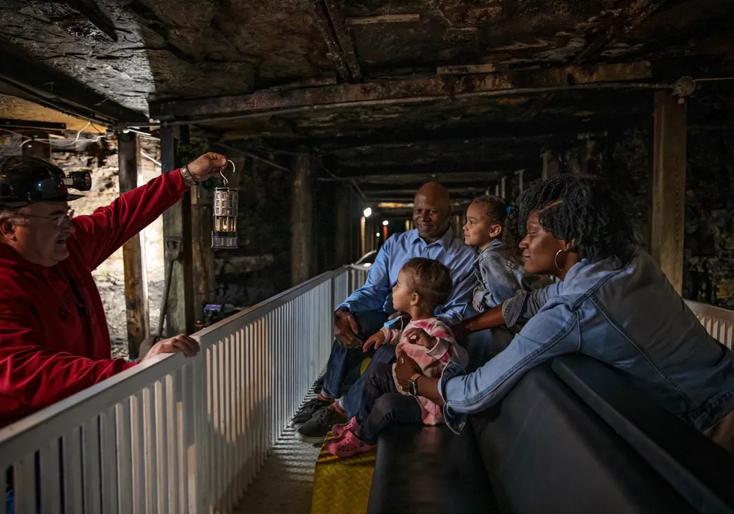 Visitors ride through the underground passages of the Beckley Exhibition Coal Mine as a guide displays a historic mining lantern. Rock walls, timber supports and low lighting reveal the working conditions once experienced by coal miners.