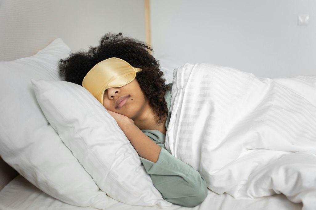 A woman sleeping under white blankets with a tan eyemask over her eyes