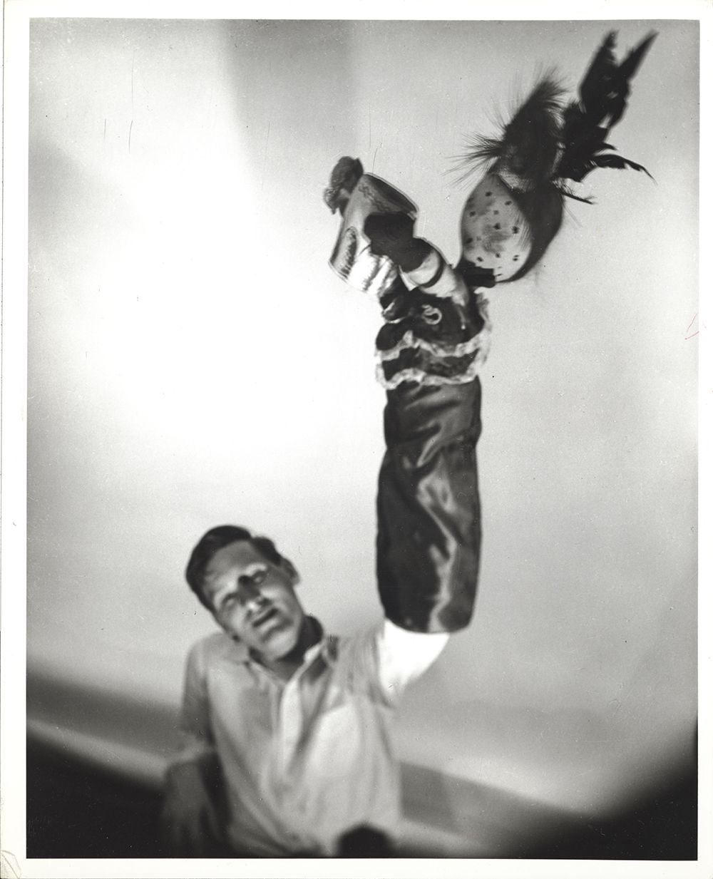 Blurry black and white photo of a man sitting in a chair wearing a white shirt and holding up a large puppet that resembles a bird.